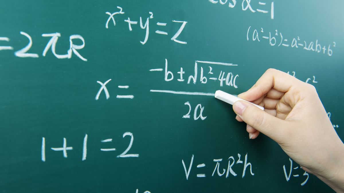 Egy kéz krétával matematikai képleteket ír egy zöld táblára.